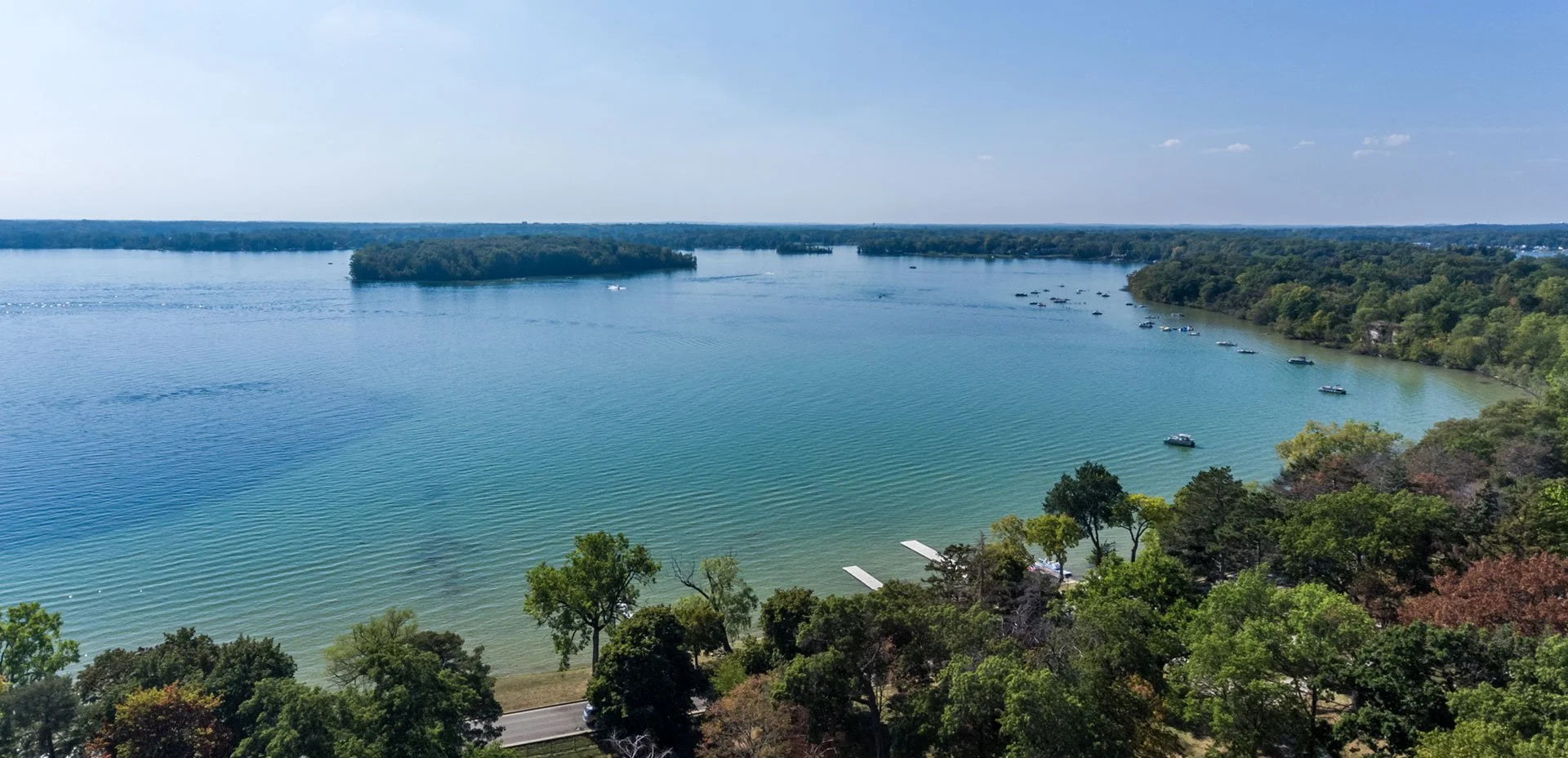 A scenic aerial view of a wide lake surrounded by dense green trees and hills, with boats docked near a small marina and a road at the shore.