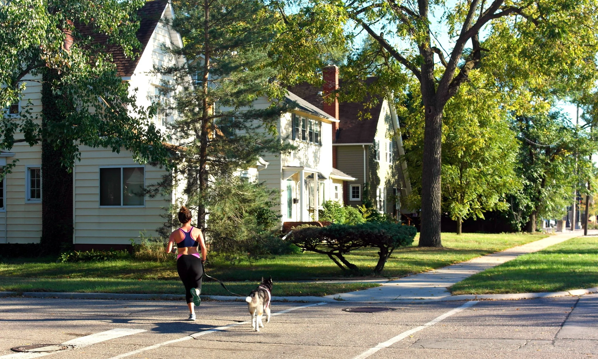 A woman jogging with a dog on a leash in a suburban neighborhood on a sunny day. Houses with trees and greenery lining the street are visible in the background.