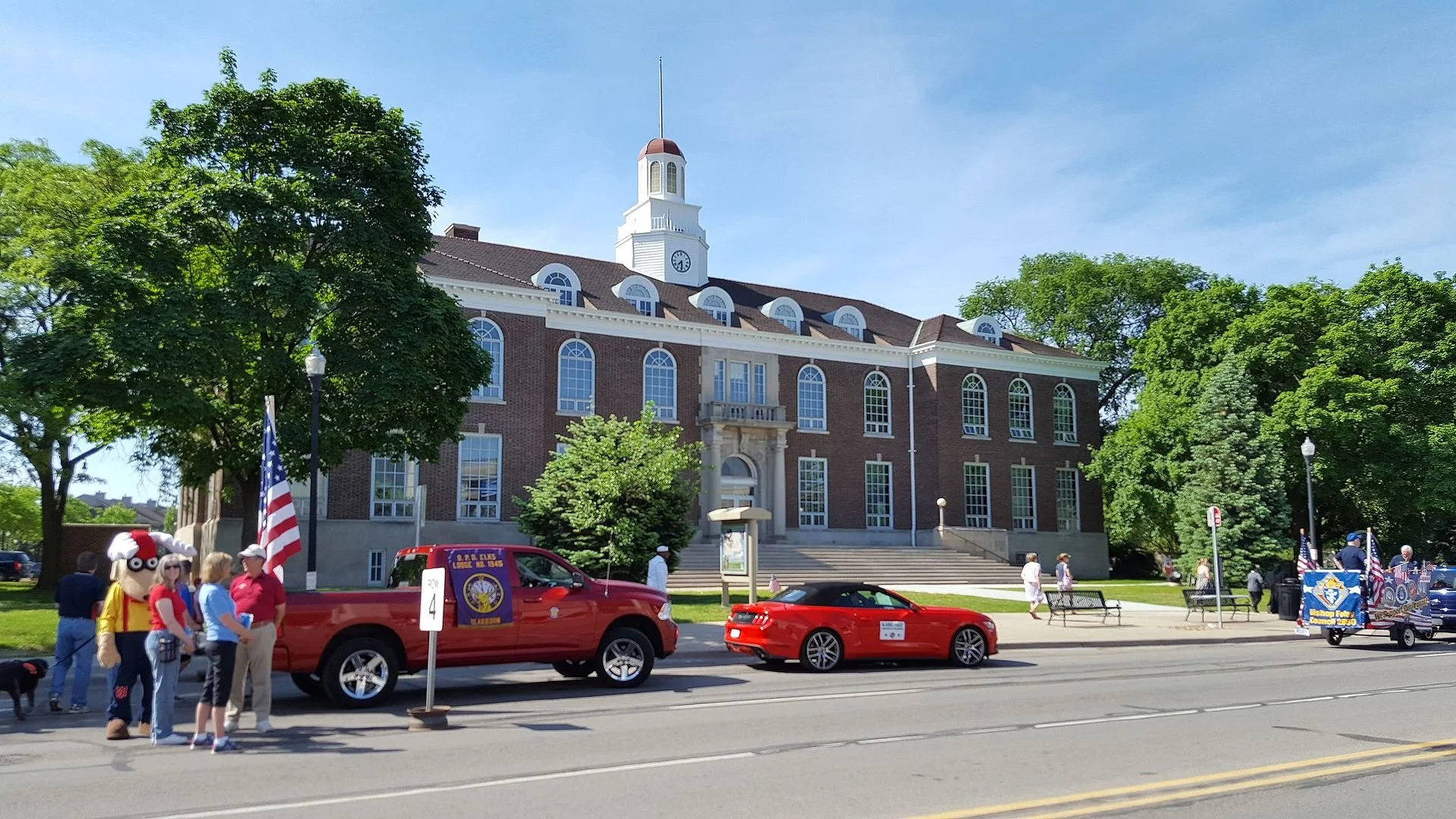 A street scene with people, vehicles, and a large brick building with arched windows and a clock tower. There are trees, American flags, and a parade or event taking place.
