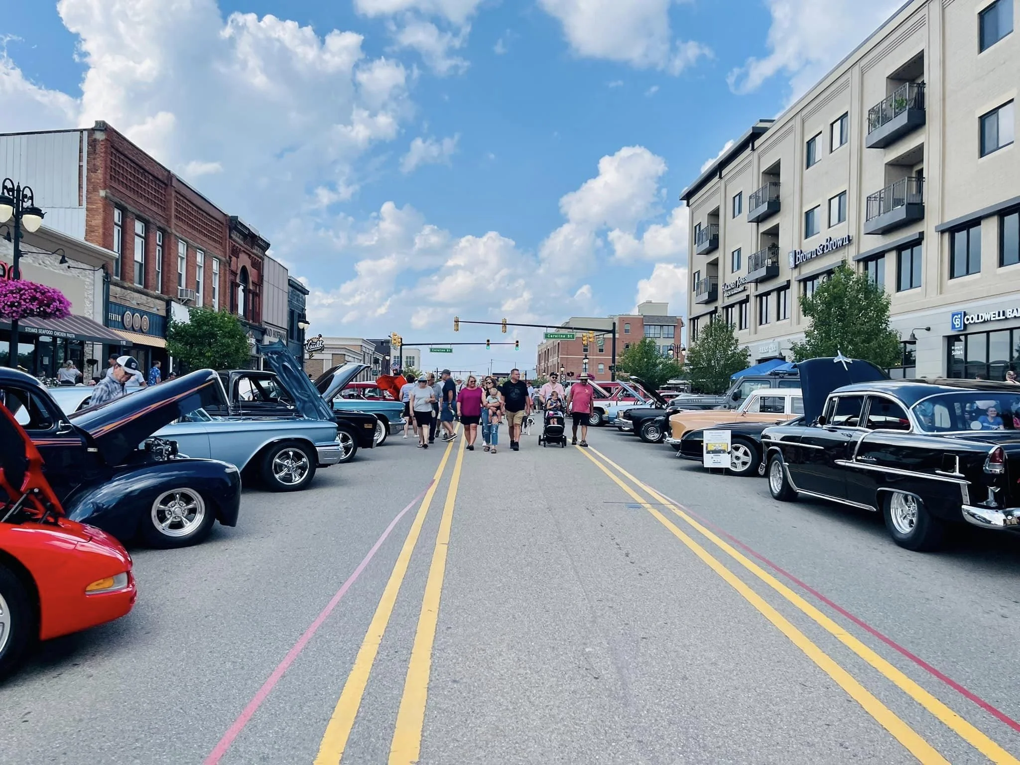 Street scene with vintage cars on display, people walking, shops in the background, blue sky with clouds.