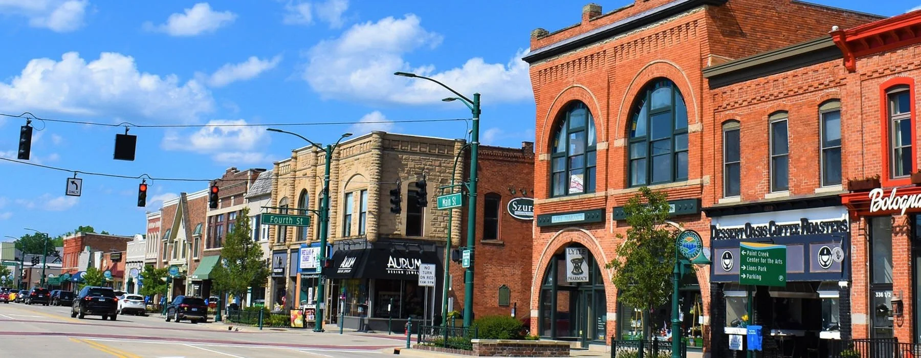 View of a downtown street with historic brick buildings, storefronts, traffic lights, and street signs for Fourth Street and Main Street on a sunny day.