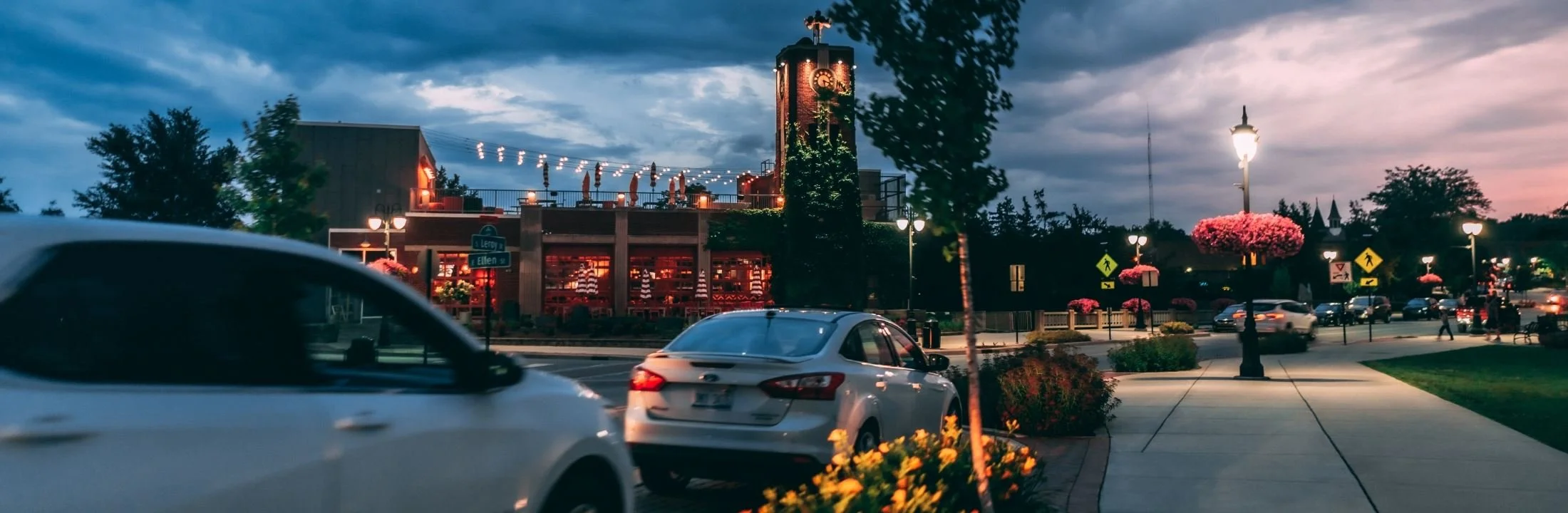 A lively town square at dusk with a clock tower, streetlights, cars, pedestrians, and flowering trees.