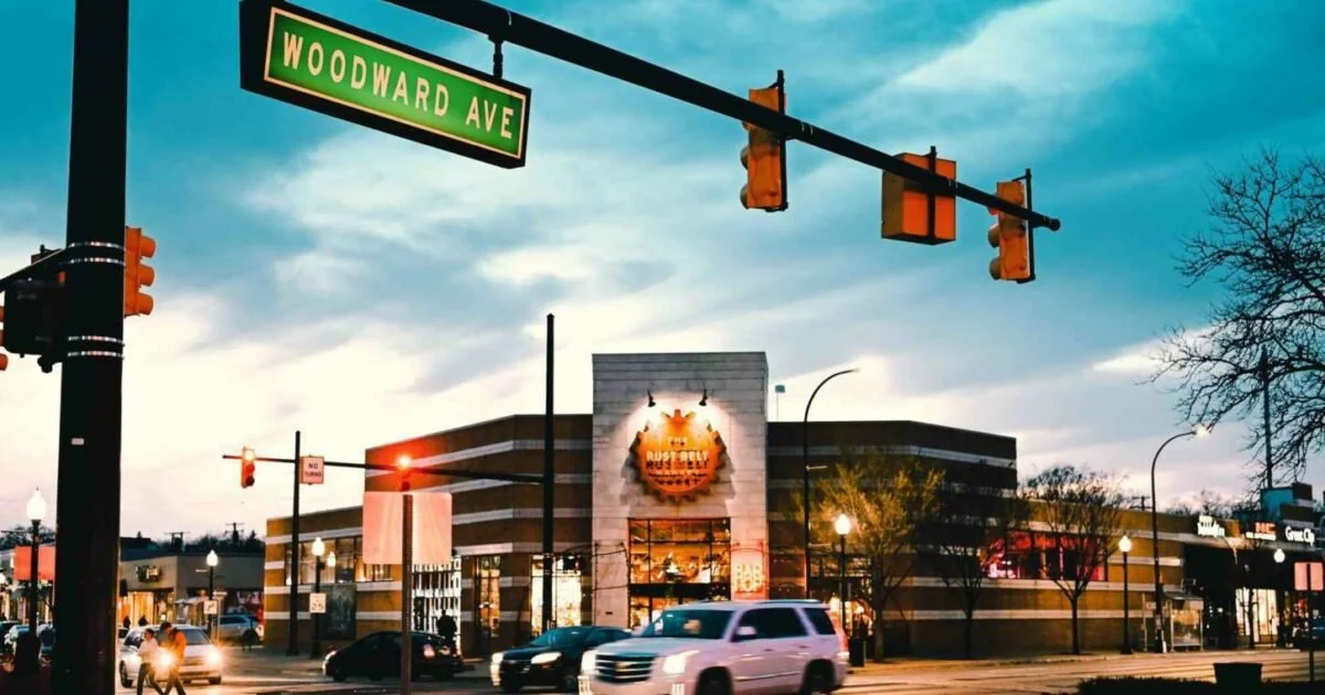 Street view of a shopping center with a sign for Woodward Ave and a building with a Bushel & Bee cafe, traffic lights, cars, and trees at dusk.