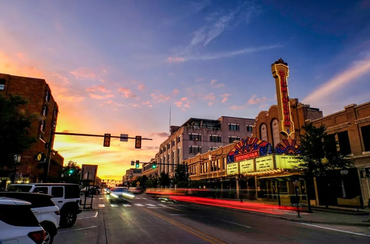 Sunset over a city street with the Birmingham theater sign illuminated, cars driving by, and buildings lining the street.