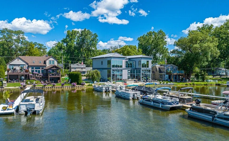 A waterfront scene with several boats docked at a marina, houses in the background surrounded by trees, and a clear blue sky with some clouds.