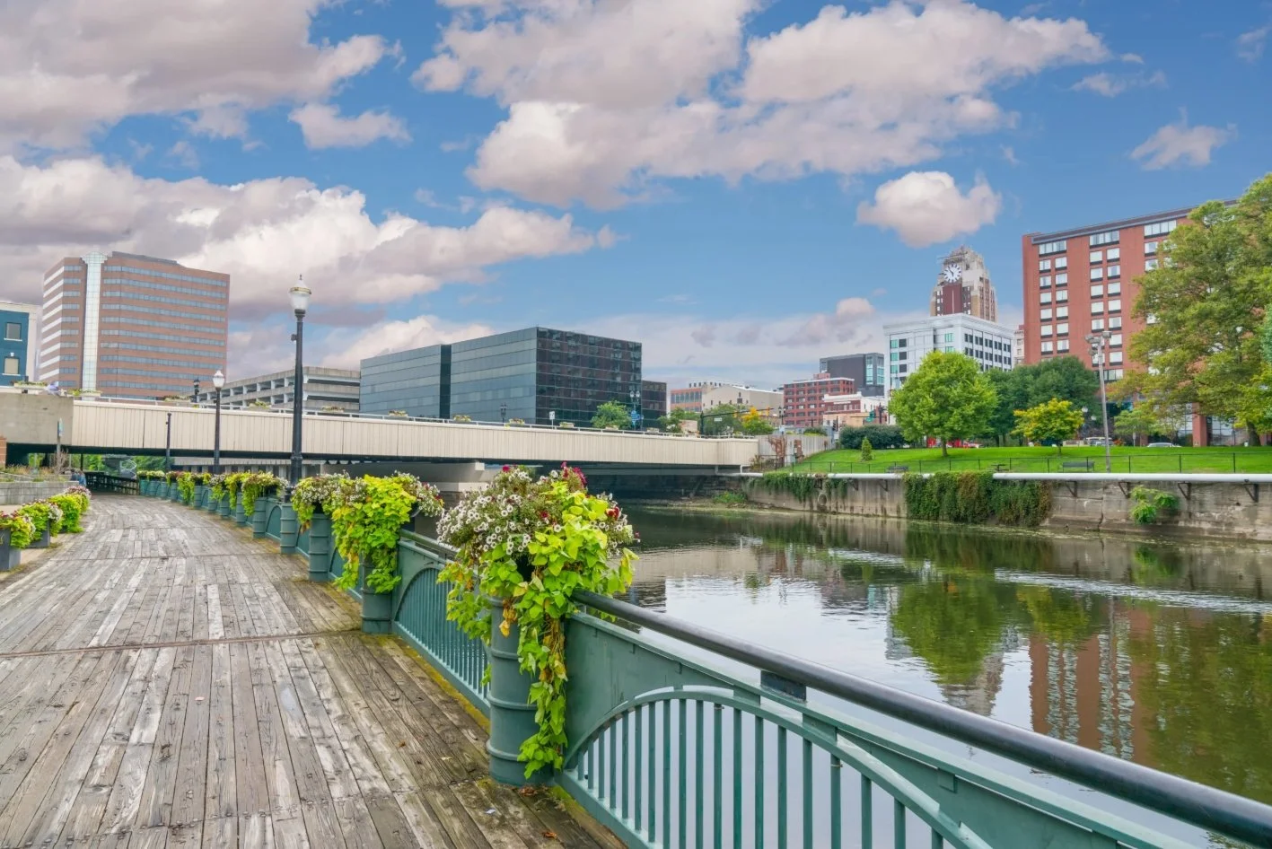 A city riverwalk scene with a wooden pathway, flower planters, lamp posts, a calm river, and modern buildings in the background under a partly cloudy sky.