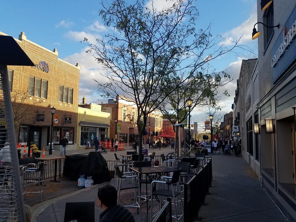 An outdoor city street scene with a large leafless tree in the center, surrounded by outdoor seating, pedestrians, storefronts, and buildings under a partly cloudy sky.