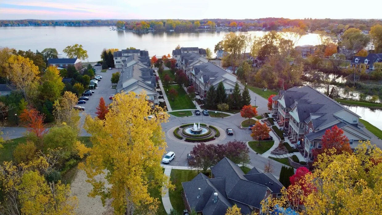 An aerial view of a residential neighborhood with multi-story townhouses, a circular driveway with a fountain in the center, and colorful autumn trees around a lake.