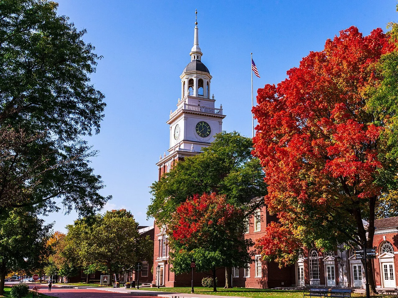 A historic clock tower on a university campus with trees showing fall foliage and a blue sky in the background.