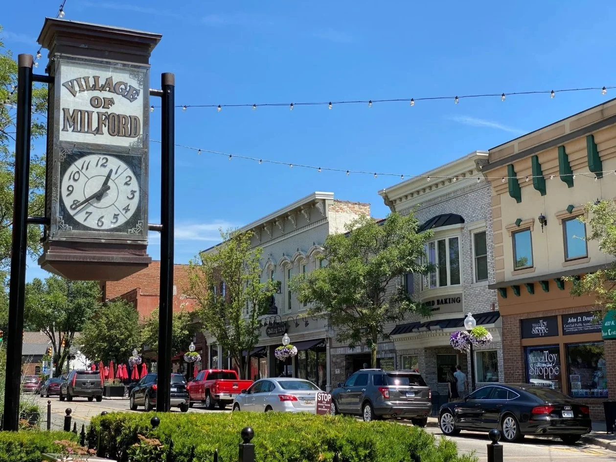 Street view of Milford with a clock tower, cars parked along the curb, storefronts, trees, and hanging string lights under a clear blue sky.