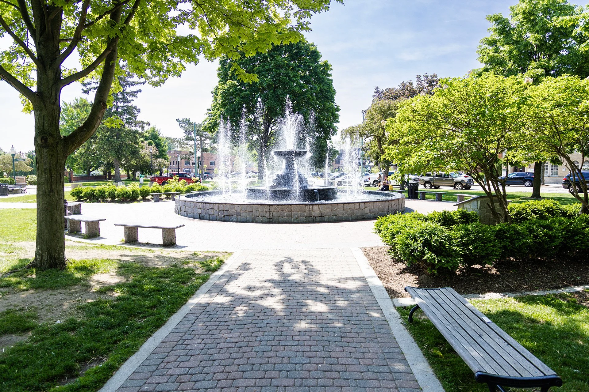 A park with a fountain in the center, surrounded by trees and benches on a sunny day.