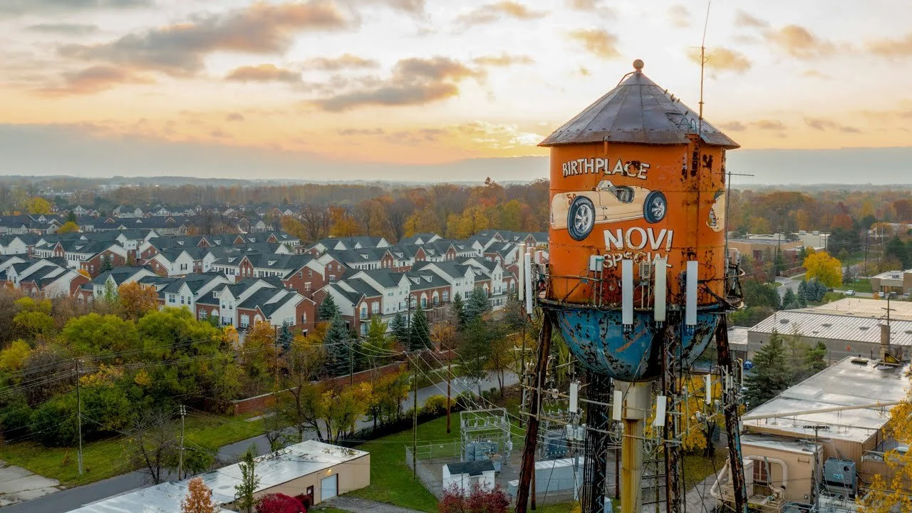 An old water tower with a painted advertisement for a birth place, Novi special, featuring a vintage car, in a suburban neighborhood during sunset with autumn foliage.