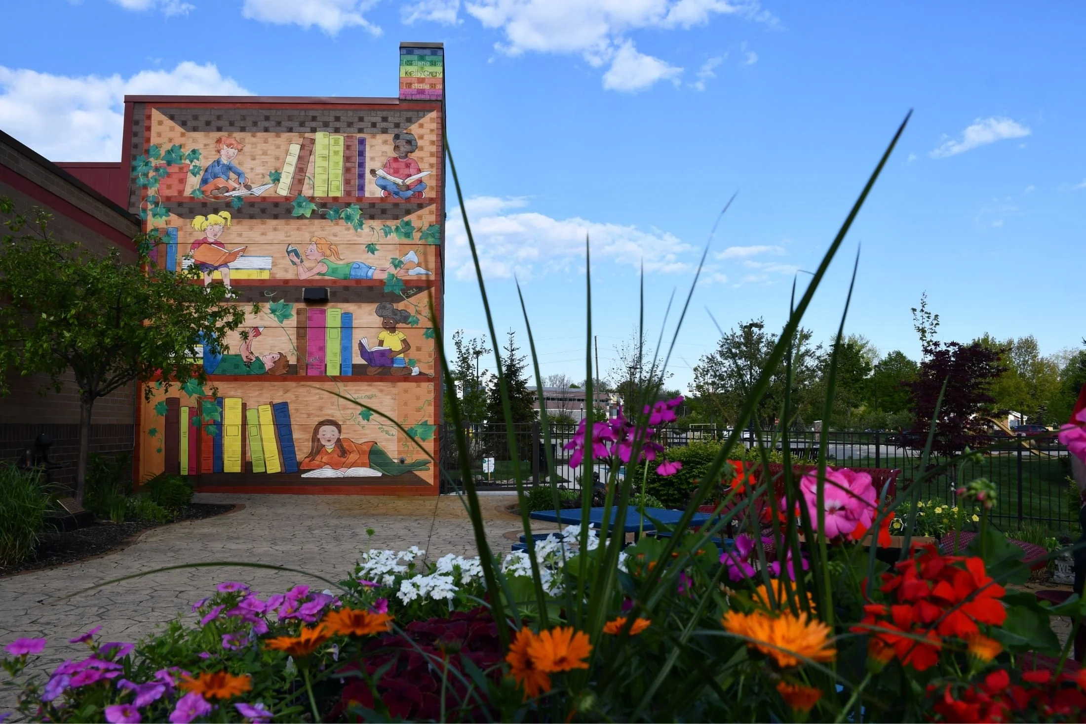 A colorful mural of children reading books painted on a building wall, with vibrant flowers and a garden in the foreground under a blue sky with scattered clouds.