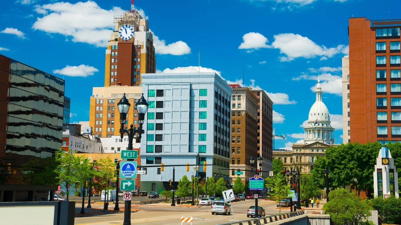 Downtown city scene with tall buildings, a street with cars, trees, and street lamps under a bright blue sky with clouds. In the background, a government building with a dome.