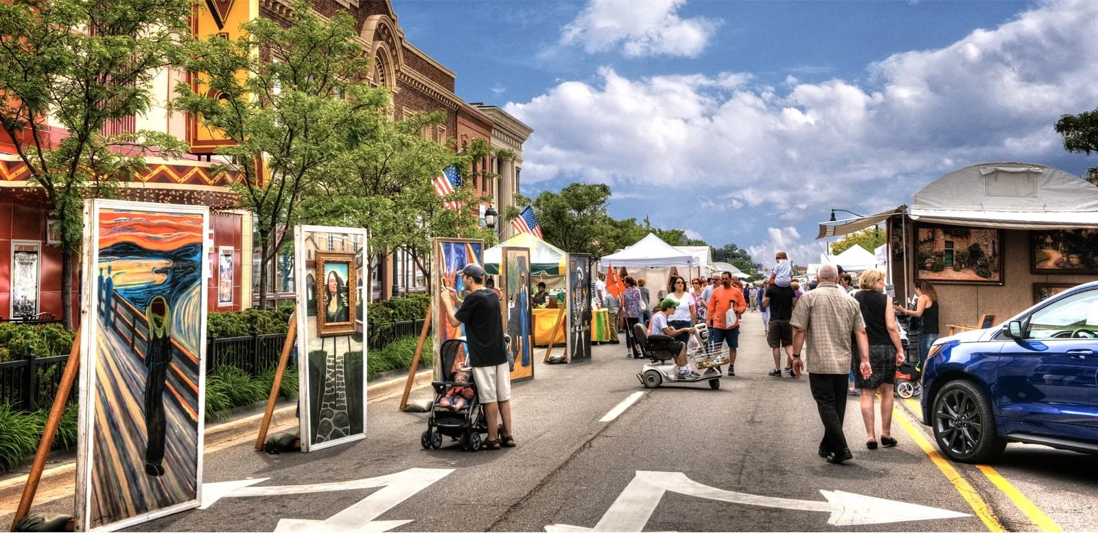 People walking and browsing art at an outdoor art festival on a city street with tents, trees, and a blue sky with clouds.