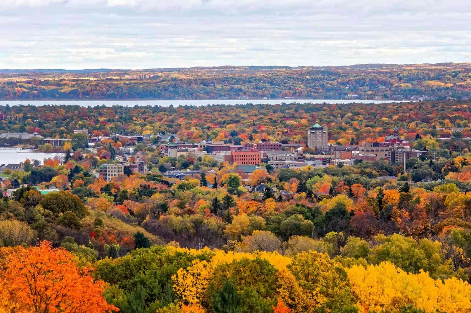 An aerial view of a city surrounded by a forest with colorful fall foliage, a river in the background, and overcast skies.