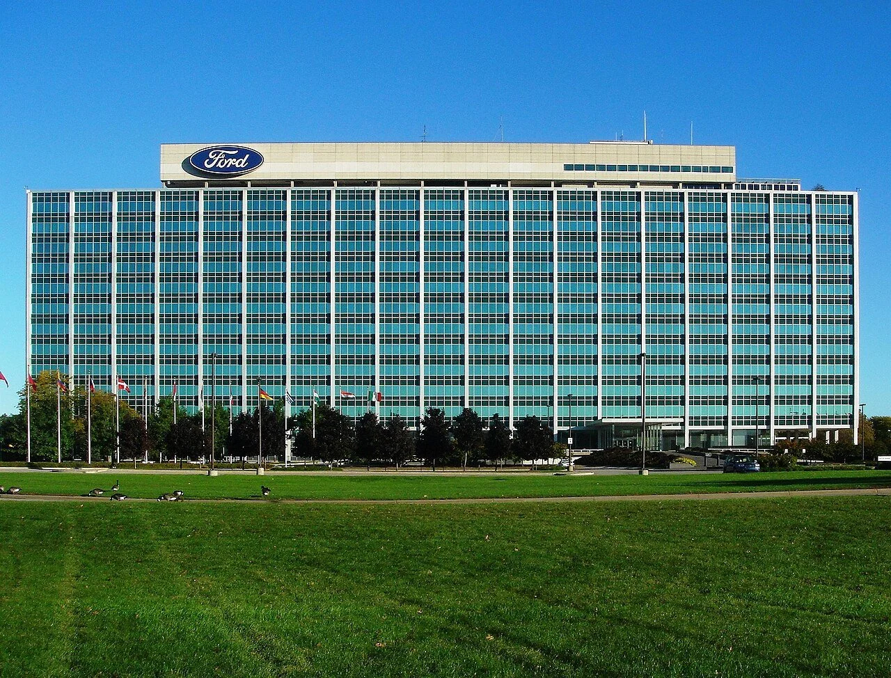 Large modern office building with Ford logo on top, surrounded by a green lawn and trees under a clear blue sky.