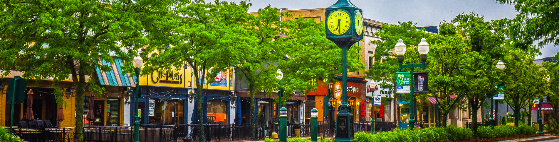 City street scene with a clock tower, green trees, storefronts, outdoor seating, and streetlights.