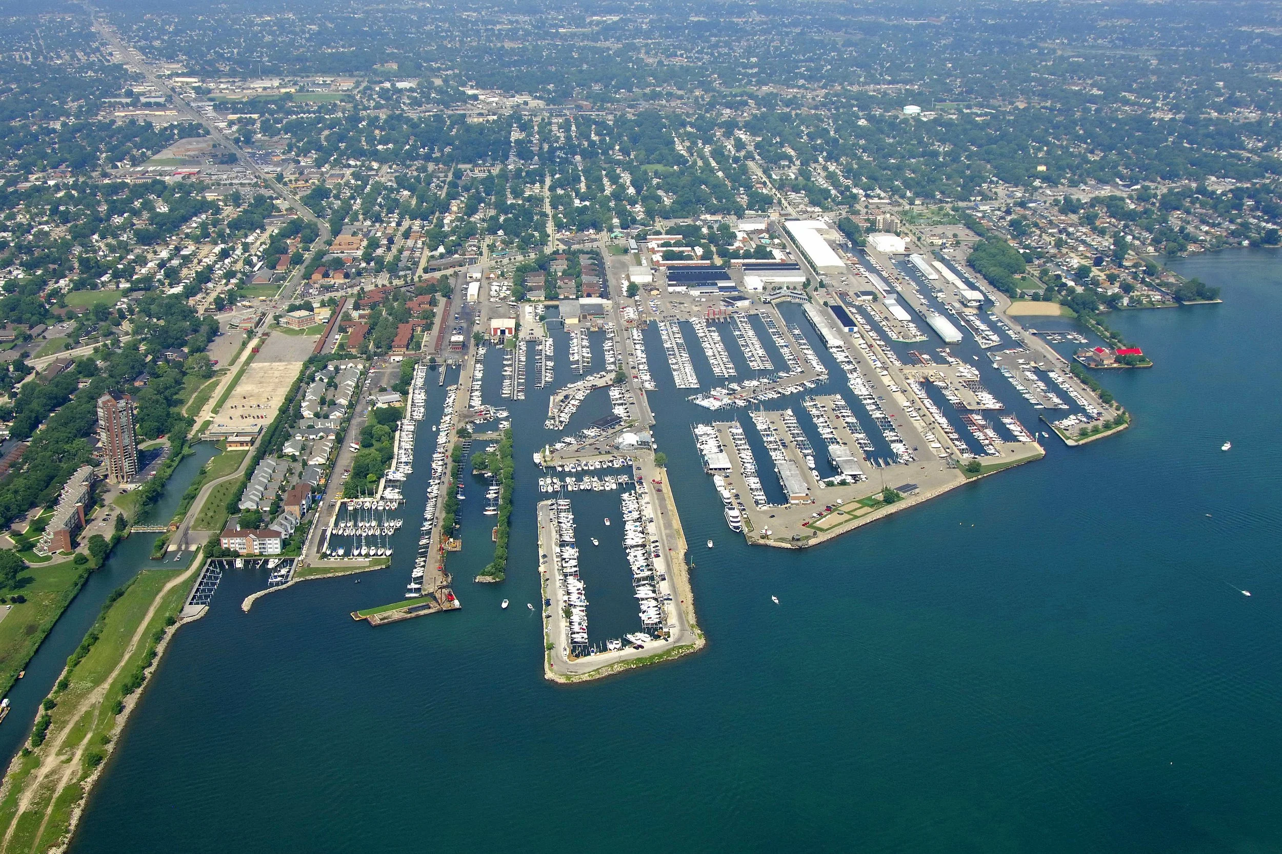 An aerial view of a harbor filled with numerous boats and yachts docked at piers, surrounded by a mix of residential and commercial buildings, with a body of water in the foreground and a cityscape extending into the background.