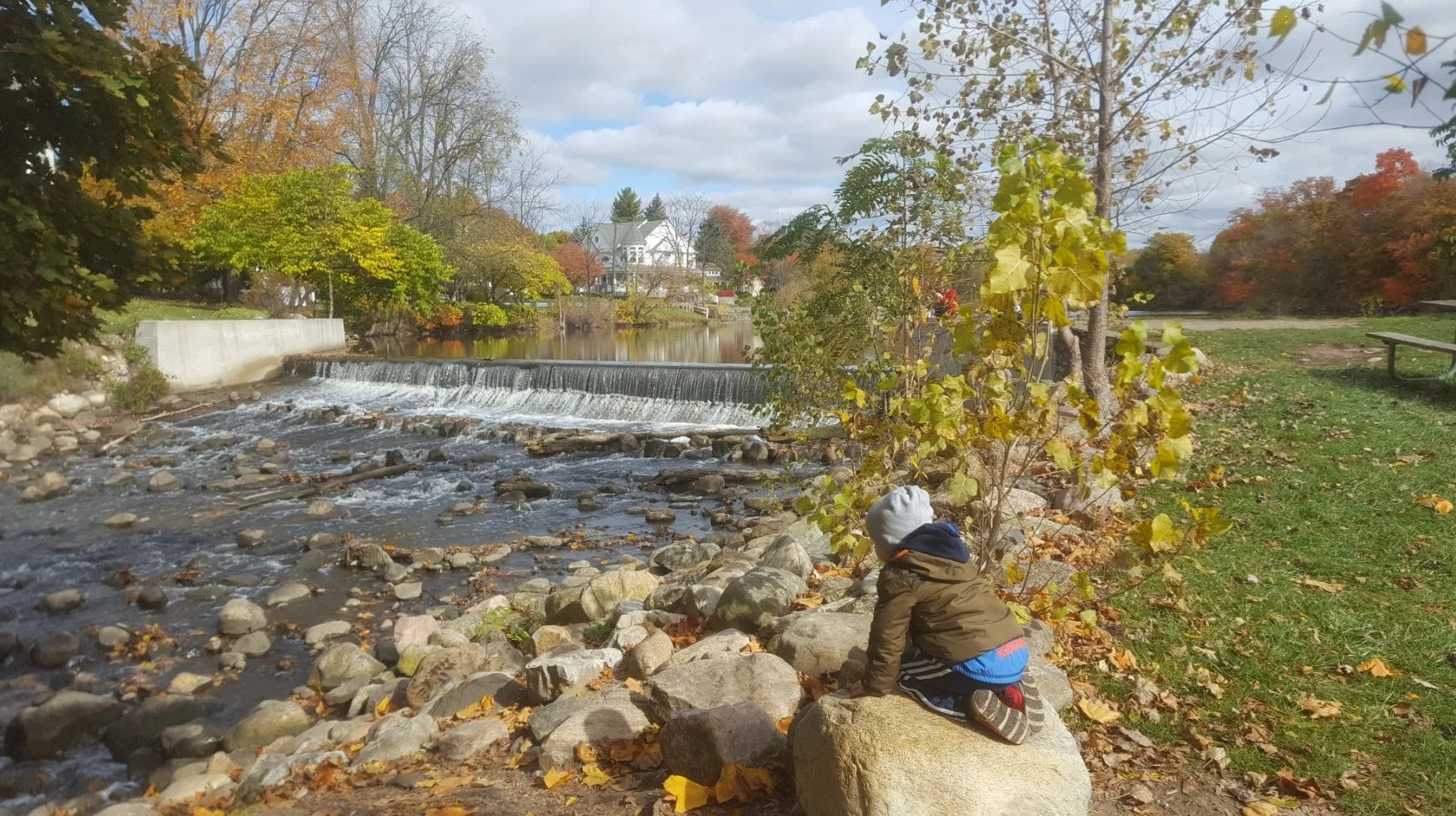 A young child wearing a coat, hat, and sneakers crouches on a rock by a river, looking at the water. The scene features autumn trees with yellow and orange leaves, a small waterfall, and houses in the background under a cloudy sky.