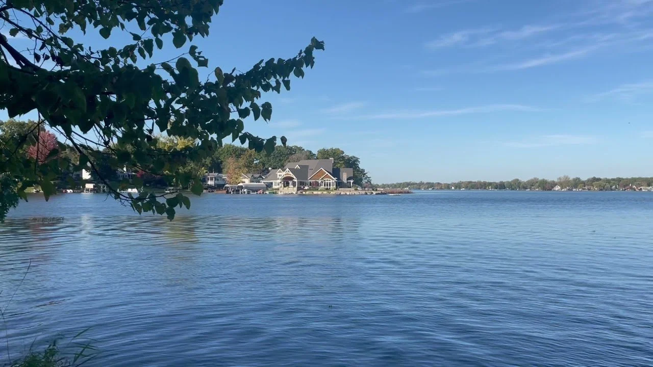 A lakeside view with a house and trees on the shore, calm water reflecting the clear blue sky, and some clouds.