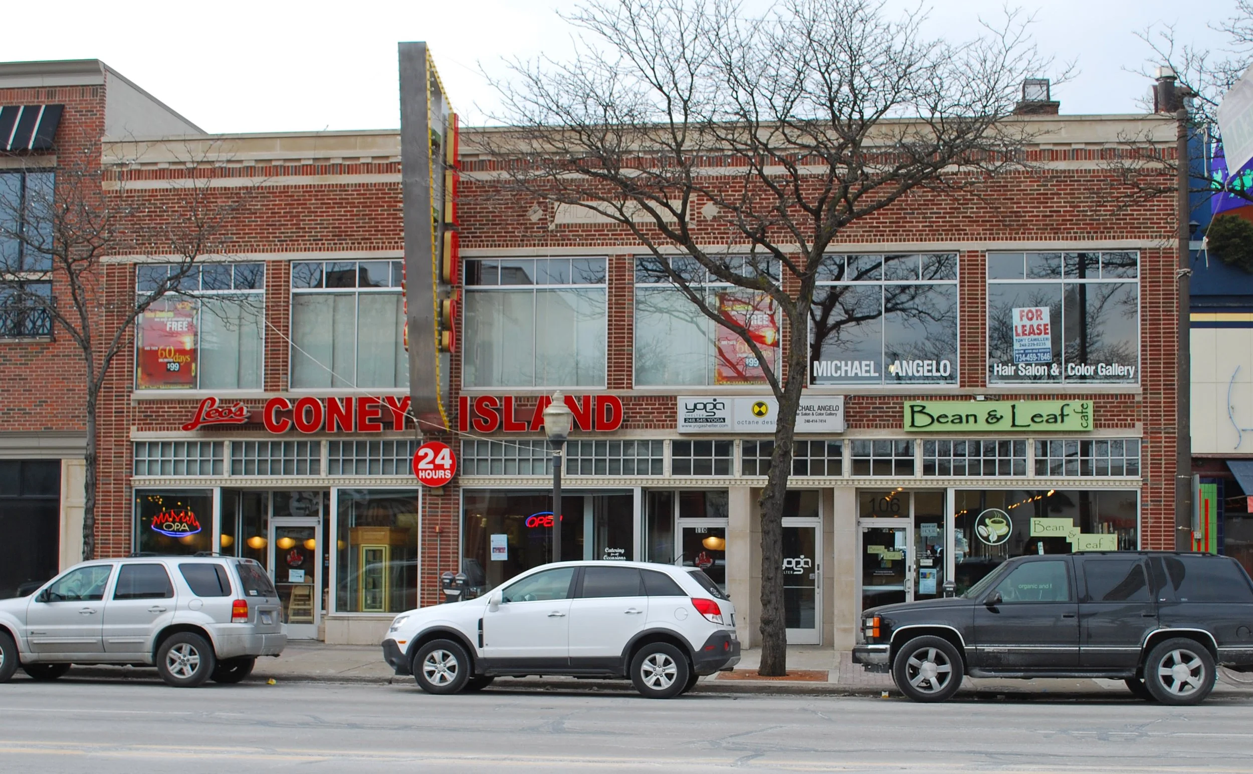A two-story brick building with various signs on the front, including a sign that says 'CONEY ISLAND' in red letters, and a '24 HOURS' sign. There are cars parked in front and leafless trees on the sidewalk.