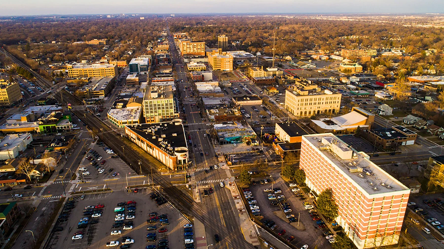 Aerial view of a downtown city area with streets, parking lots, and various buildings, some with rooftops, under a clear sky.