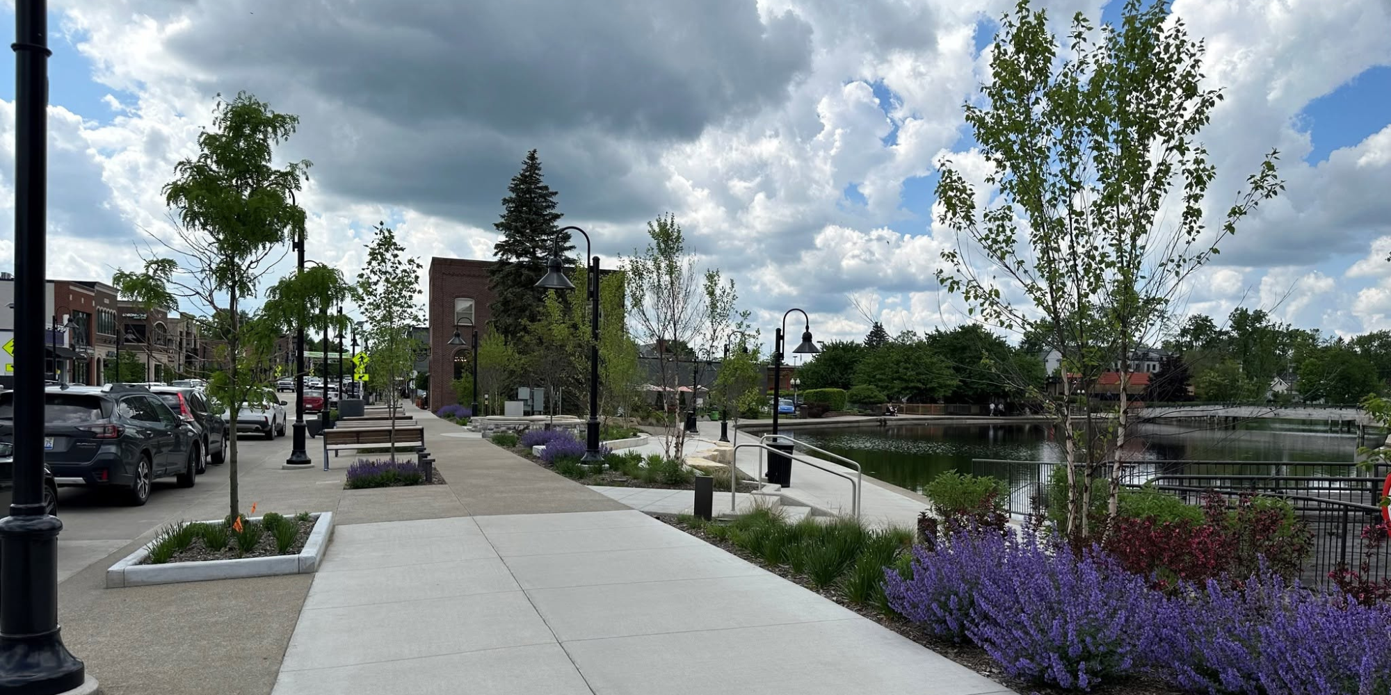 A sidewalk along a parking lot with parked cars and trees, overlooking a small pond with railings, benches, and greenery under a cloudy sky.