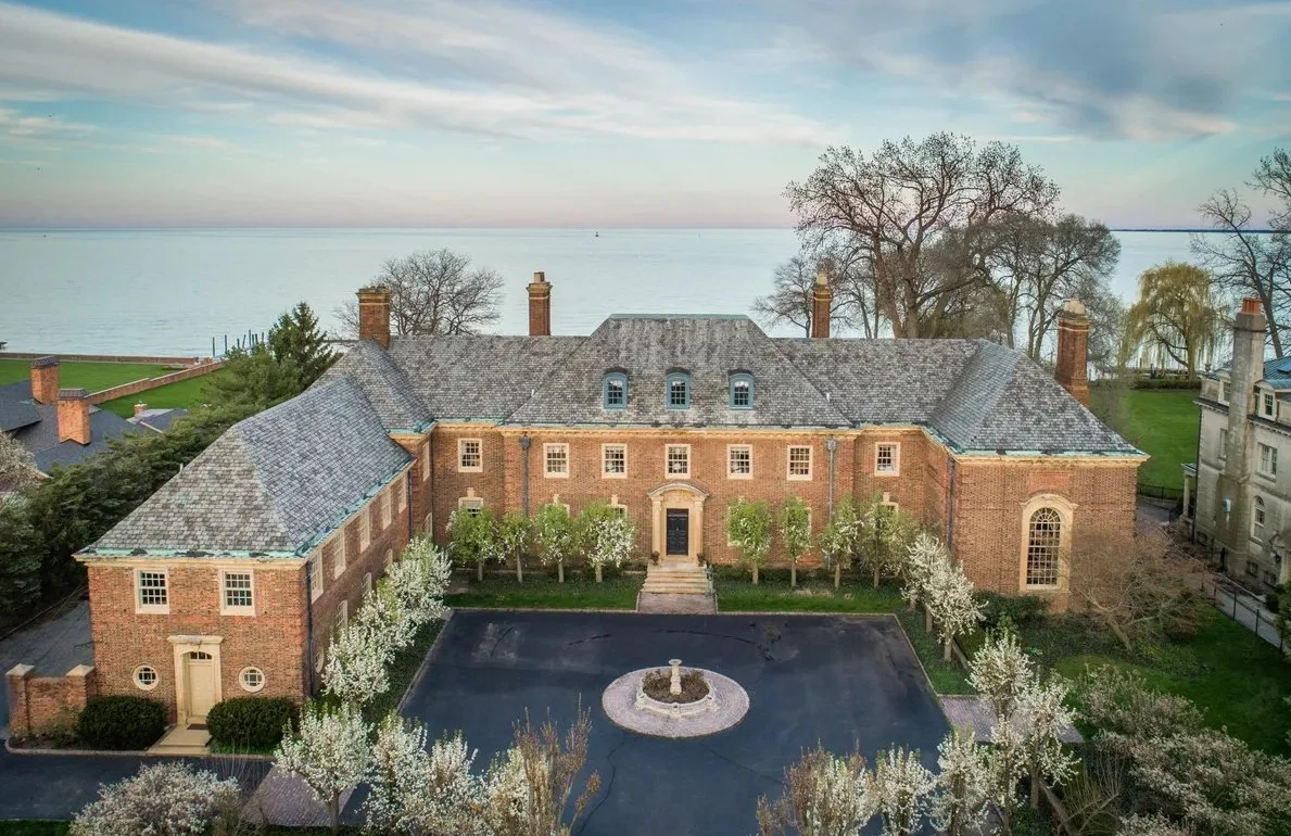 A large brick mansion overlooking a body of water, with a circular fountain in front and blooming trees around it.