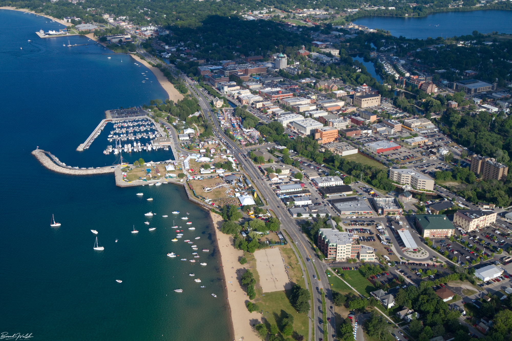 Aerial view of a coastal city with a marina containing sailboats, a beach, a busy downtown area with shops and buildings, and green trees along the roads and in the background.