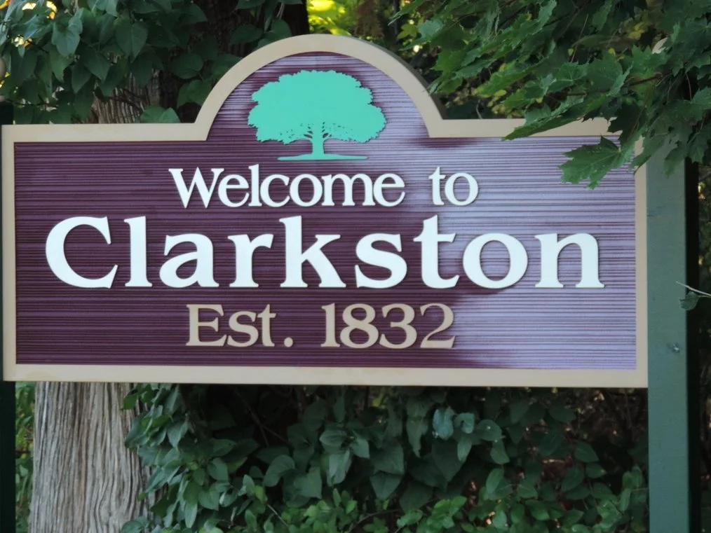 Welcome sign for Clarkston with a tree graphic, established in 1832, surrounded by greenery.
