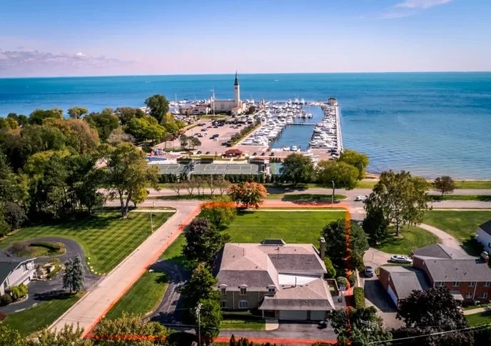Aerial view of a coastal city with a marina, harbor, and lighthouse, surrounded by green parks and residential houses.