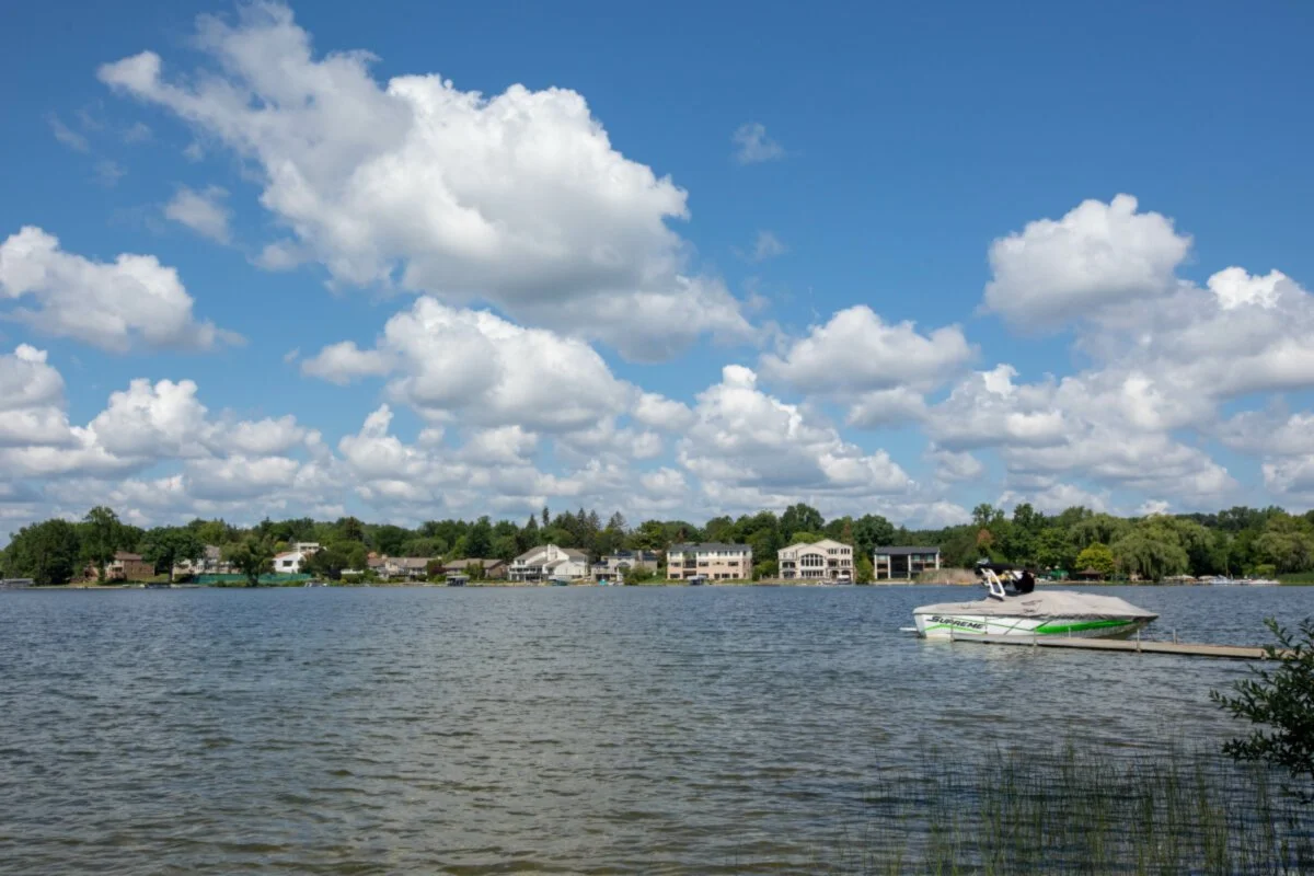 A lake with houses and trees along the shoreline, a speedboat docked at a pier, and a partly cloudy sky.