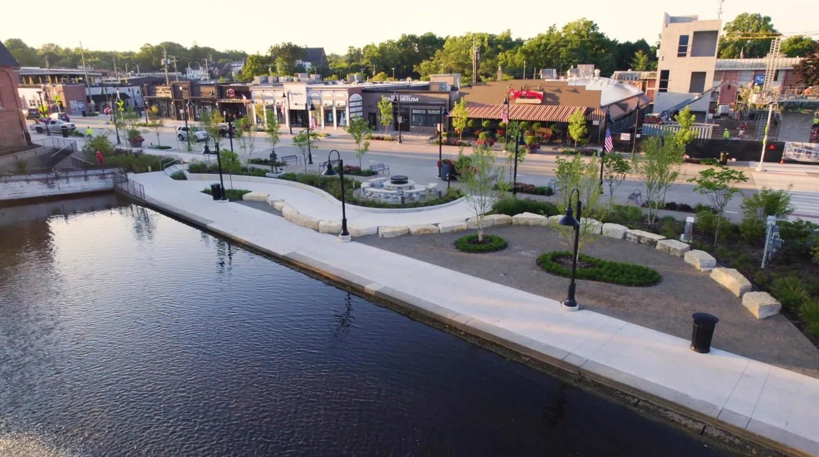 A small park area along a river with trees, benches, street lamps, and a fire pit, in front of a busy shopping street with storefronts and buildings, under a clear sky.
