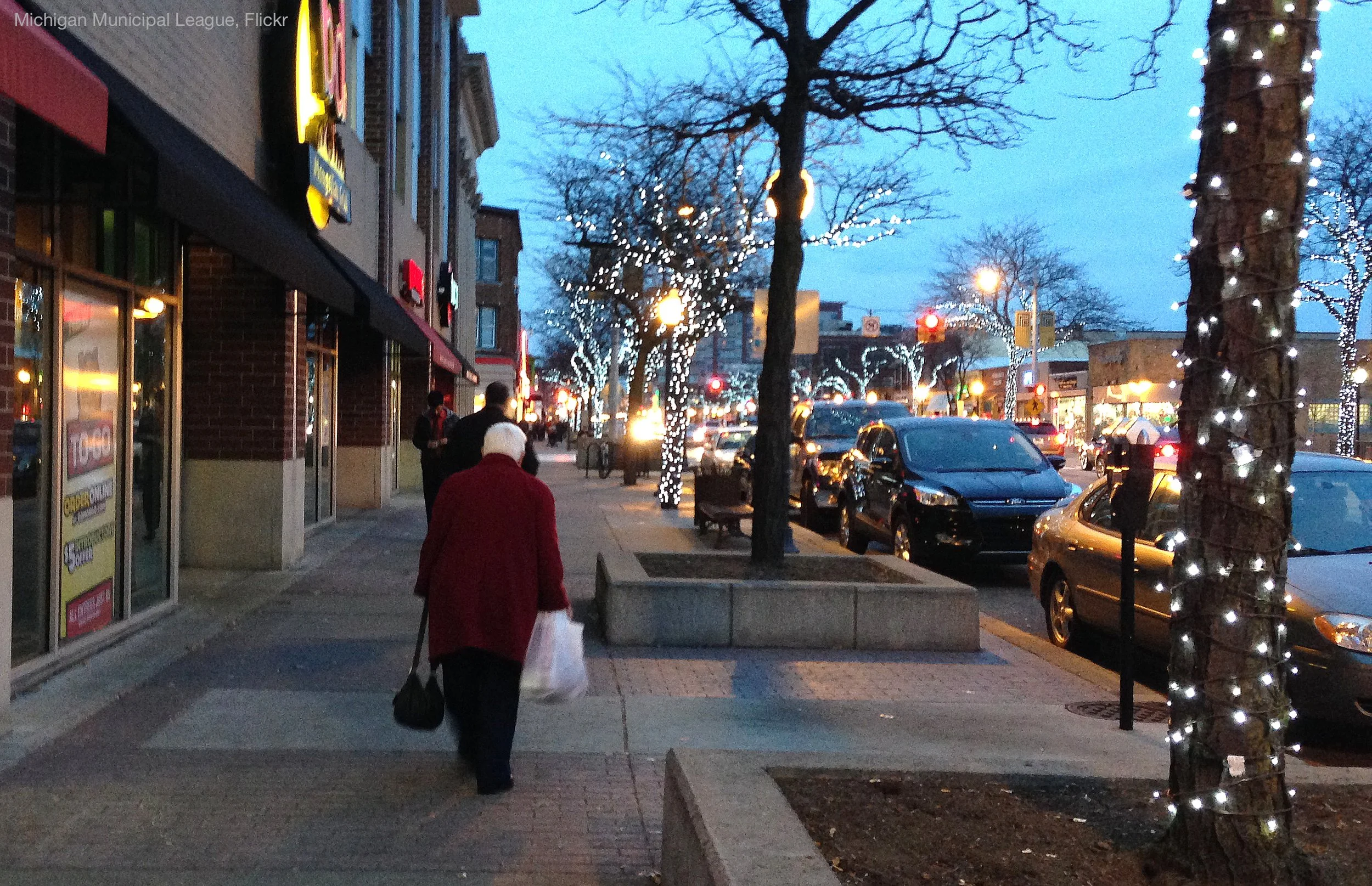 A city sidewalk during evening with people walking, lined with trees wrapped in string lights. Cars are parked along the street, and buildings with store signs are visible on the left. One person wears a red coat and carrying a shopping bag, walking