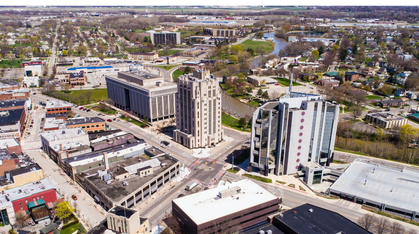 An aerial view of a downtown area with tall office buildings, smaller commercial and residential buildings, and a river running through a park. Roads and parking lots are visible.