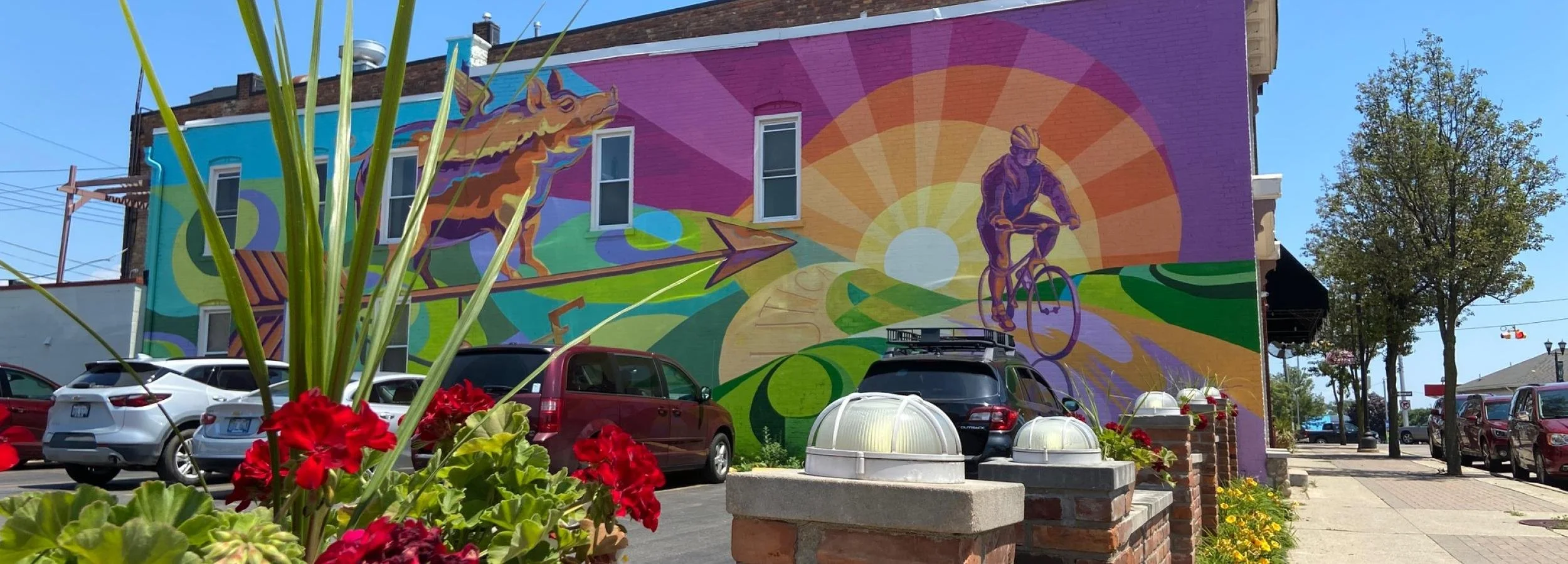 Colorful mural on a building depicting a unicorn, a cyclist, and a sunset landscape with vibrant colors, with parked cars and a sidewalk in the foreground.