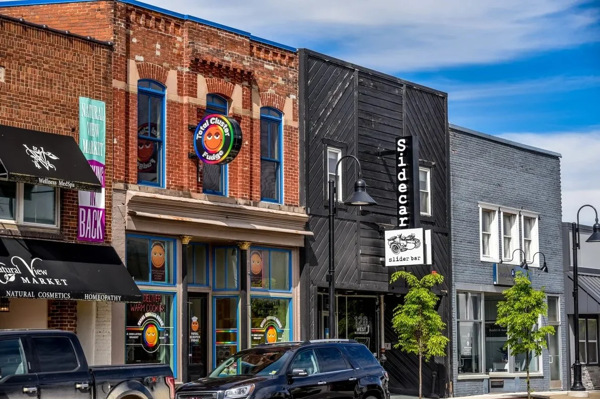 Street view of small businesses including a natural wellness market, a slide bar, and a store called Total Cluster Fudge, with brick and painted facades, trees, parked cars, and street lamps under a partly cloudy sky.