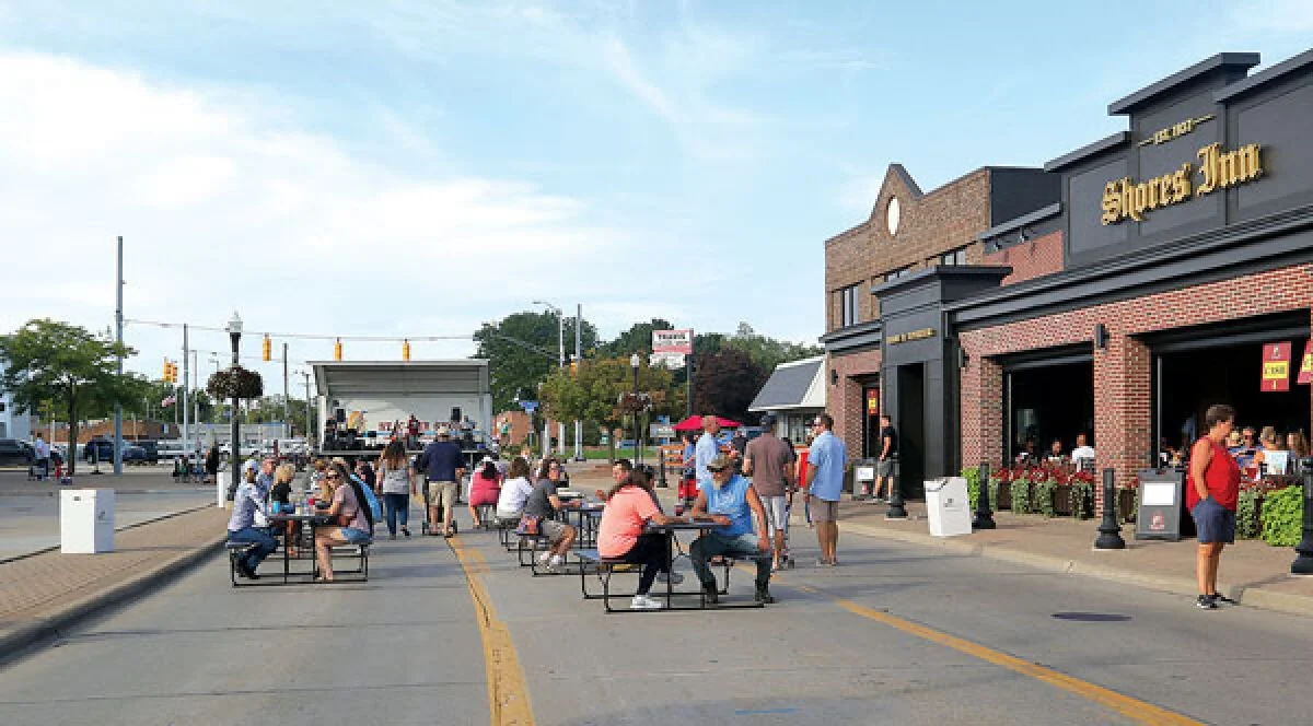 People sitting at outdoor tables near a food establishment called Shoes Inn, with a stage and concert setup in the background on a city street.