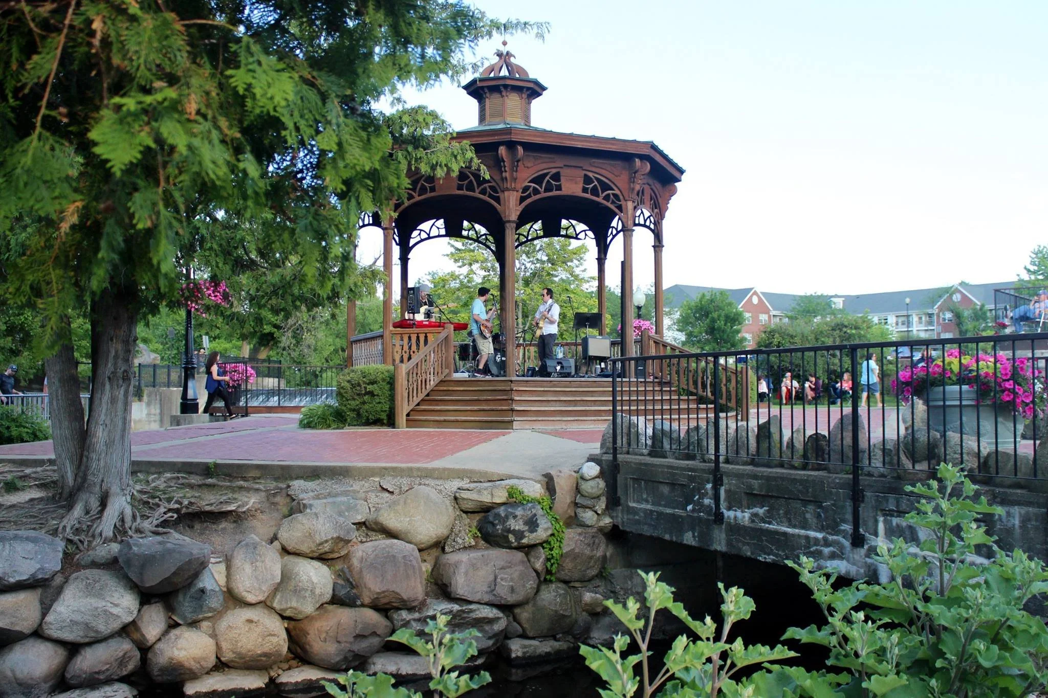 A band performs on a gazebo stage in a park, surrounded by trees, pink flowers, benches, and people walking and sitting nearby.