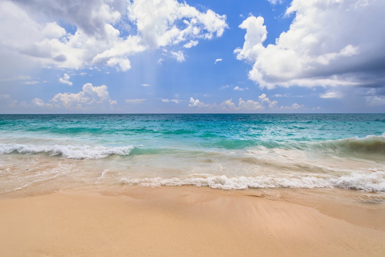 Sunny beach with white sand, turquoise ocean waves, and a partly cloudy blue sky.