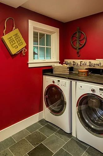 Laundry room with red walls, a white window, a yellow 'Need Help' sign, a decorative wall piece, and a countertop with laundry supplies, housing a washing machine and dryer.