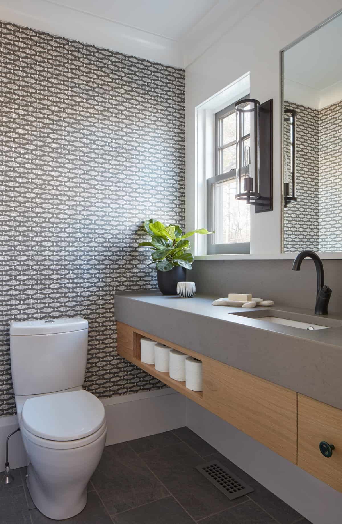 Modern bathroom with patterned black and white fish-scale wallpaper, a white toilet, a gray countertop with a built-in sink, a black faucet, a mirror, a black wall-mounted light fixture, a window, a potted plant, and shelves with toilet paper rolls.