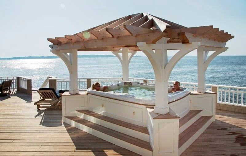 Two people relaxing in a hot tub on a wooden deck by the ocean, under a wooden gazebo with a view of the water.