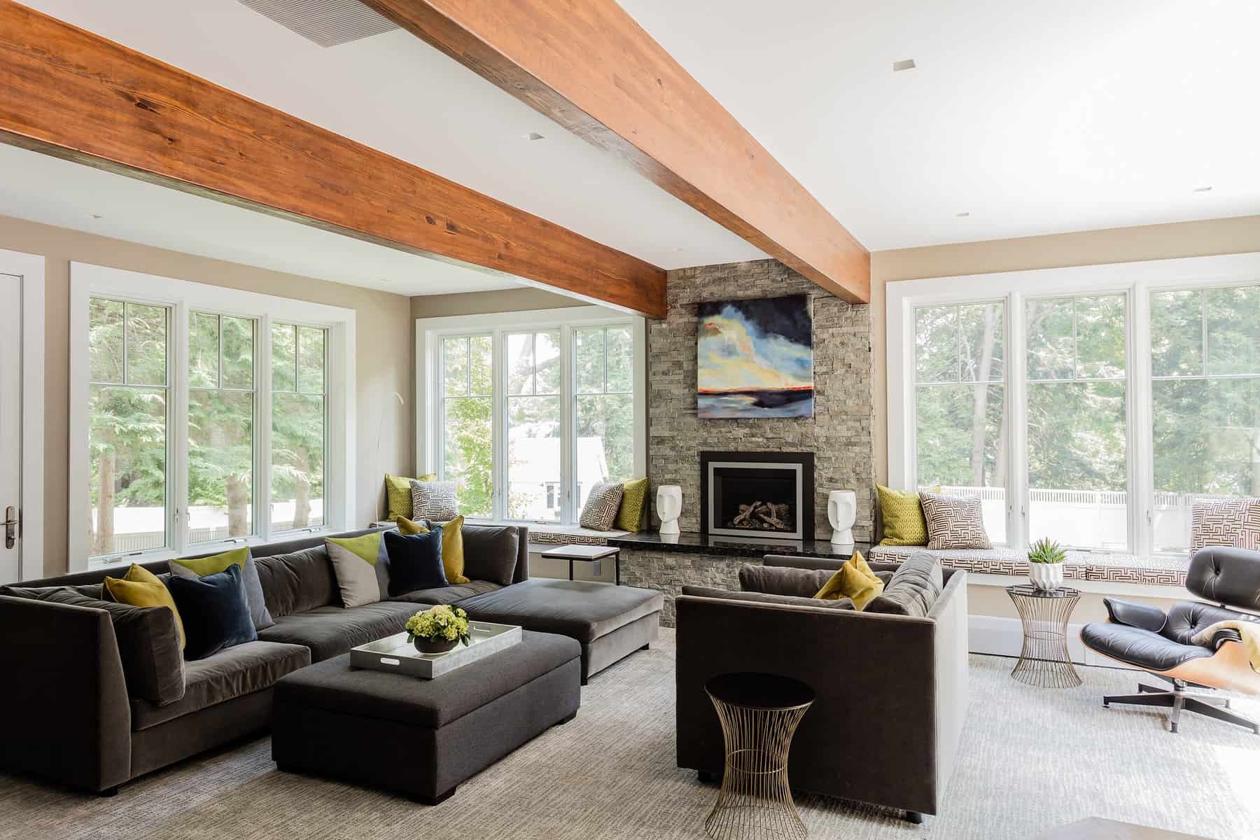 A spacious living room with large windows, a stone fireplace with a colorful painting above, and a ceiling with wooden beams. The room is decorated with dark sofas, accent pillows, side tables, and a potted plant, with natural light streaming in from