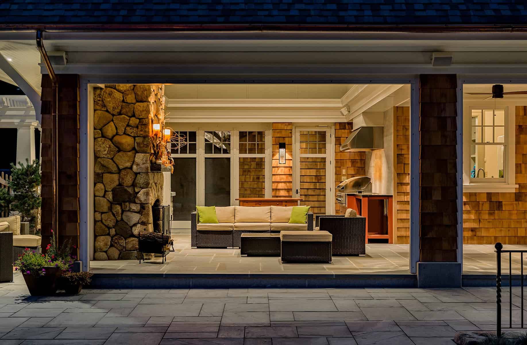 View of a cozy outdoor patio with seating, a stone fireplace, and a kitchen under a porch at night. There are green cushions on the sofa, a potted plant, and warm lighting inside.