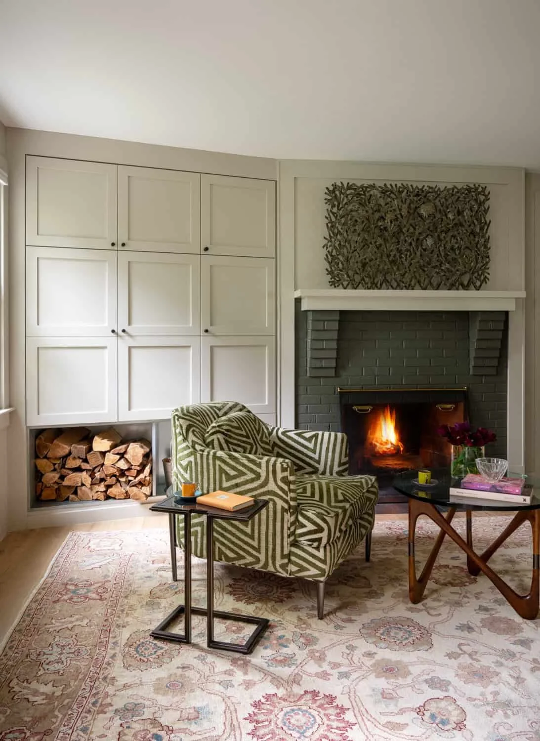 Living room with a fireplace, patterned armchair, side tables, and wood logs by the wall.