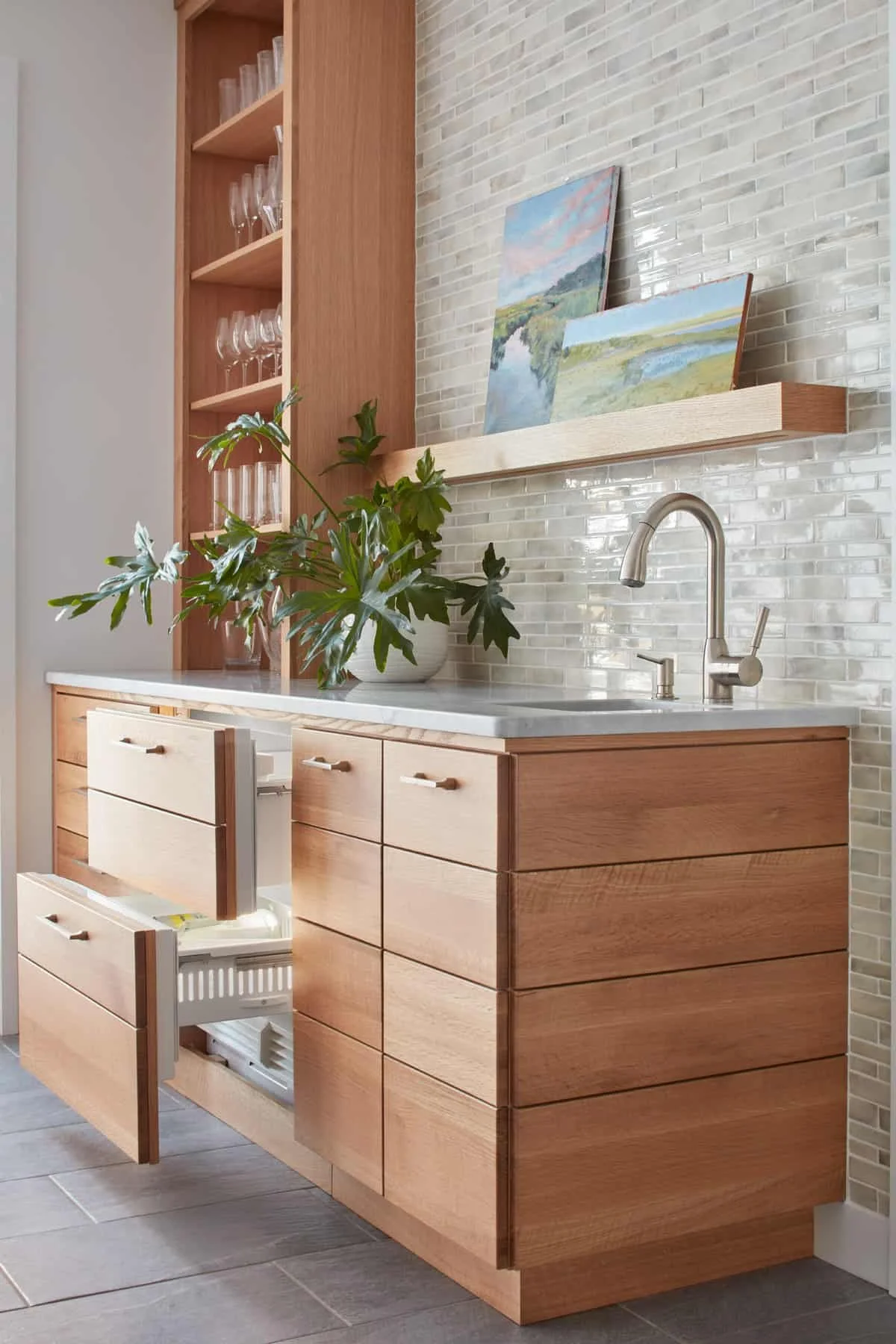 Kitchen with wooden cabinets, a white marble countertop, a metal faucet, a potted green plant, and two landscape paintings on a shelf above the sink, with a white tiled backsplash and gray floor tiles.