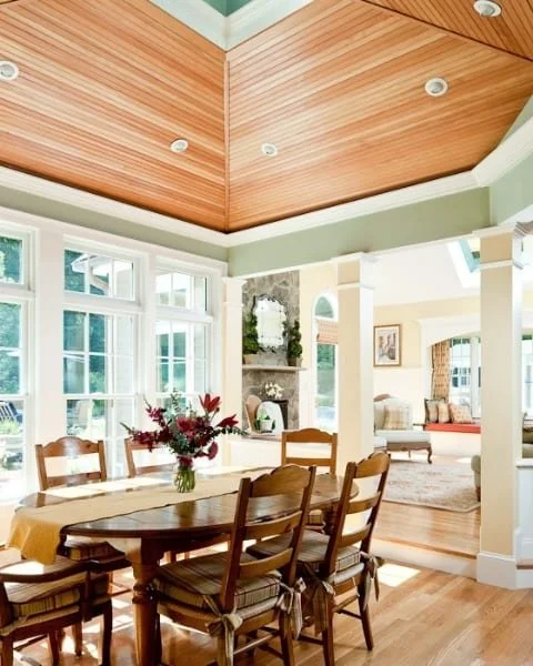 Bright dining room with a wooden table, six chairs, and a floral centerpiece. Sunlight streams through large windows. The room connects to a living area with a fireplace and cozy seating.