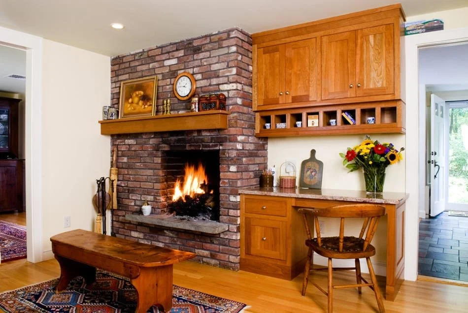 Living room with a brick fireplace, wooden cabinetry, a small wooden bench, and a vase of colorful flowers.
